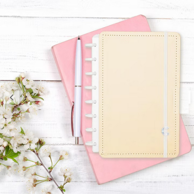 Dois cadernos rosa e bege com caneta branca e flores brancas sobre madeira branca