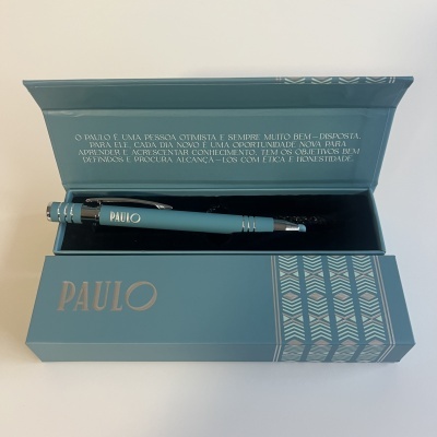 Caneta esferográfica azul dentro de caixa azul com padrão geométrico e texto em português no interior da tampa.