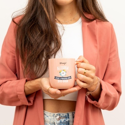 Caneca cor de rosa clara com texto e ilustração, segurada por mulher com casaco rosa