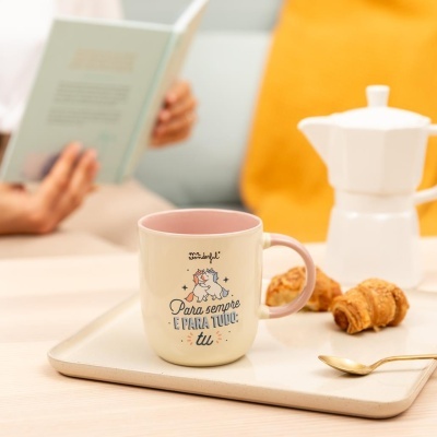 Caneca cerâmica branca com interior e asa cor-de-rosa com desenho de dois unicórnios e texto em português, numa bandeja com croissants, colher dourada e cafeteira branca, em ambiente acolhedor.