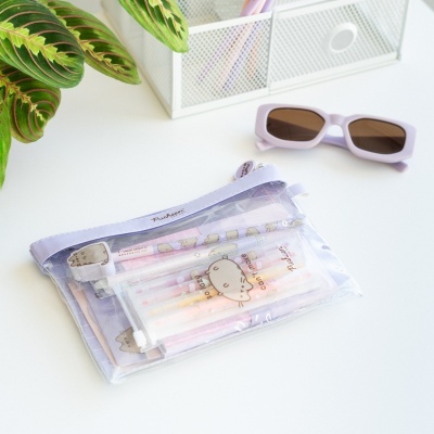 Estojo transparente lilás com artigos de papelaria com desenho do gato Pusheen, óculos de sol lilás e planta verde num fundo branco.