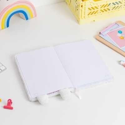 Caderno felpudo branco aberto sobre mesa branca com objetos de escritório e decoração coloridos