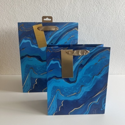 Duas sacolas de papel azuis com padrão de mármore e detalhes dourados, com alças de fita dourada.