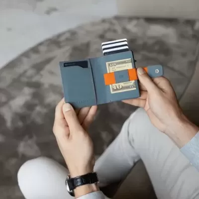 Carteira azul escura aberta com dinheiro e cartão, nas mãos de uma pessoa sentada num sofá cinzento.