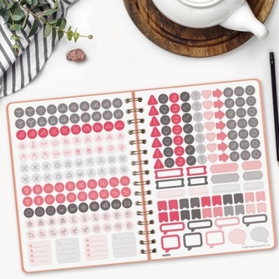 Caderno com adesivos coloridos variados em mesa com guardanapo, pedra, caneta e bule