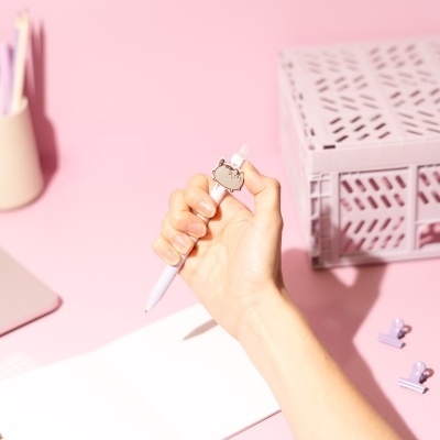 Caneta branca com decoração Pusheen numa mão, mesa rosa com acessórios de escritório.