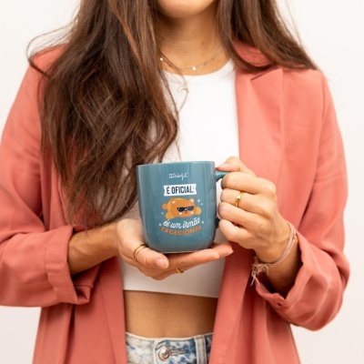 Caneca azul com texto e ilustração de urso sendo segurada por mulher com casaco rosa.