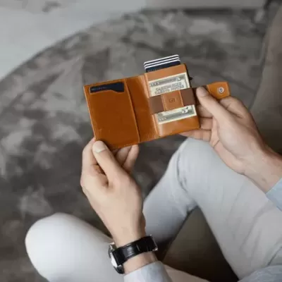 Carteira de pele castanha com cartões, nas mãos de pessoa sentada num sofá cinza.