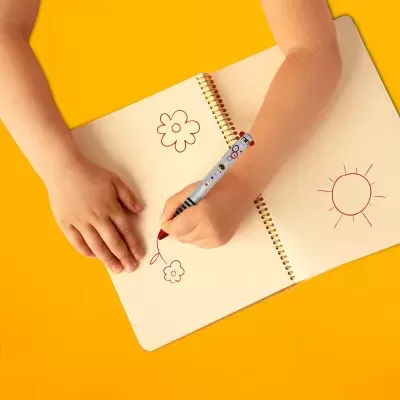Criança a desenhar flores e sol num caderno com caneta decorada sobre fundo amarelo.