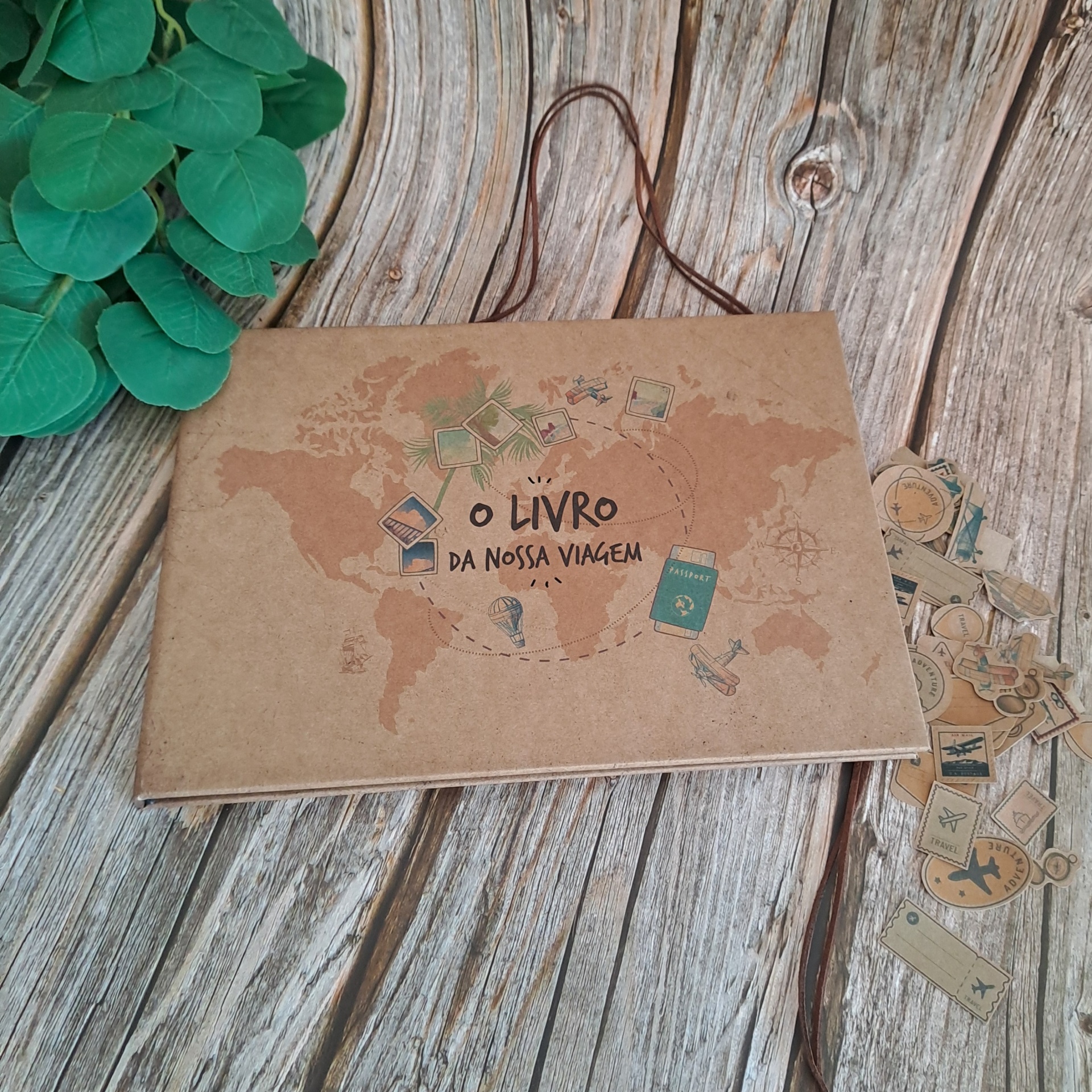 Livro 'O LIVRO DA NOSSA VIAGEM' com capa castanha e mapas, sobre madeira com folhas verdes.