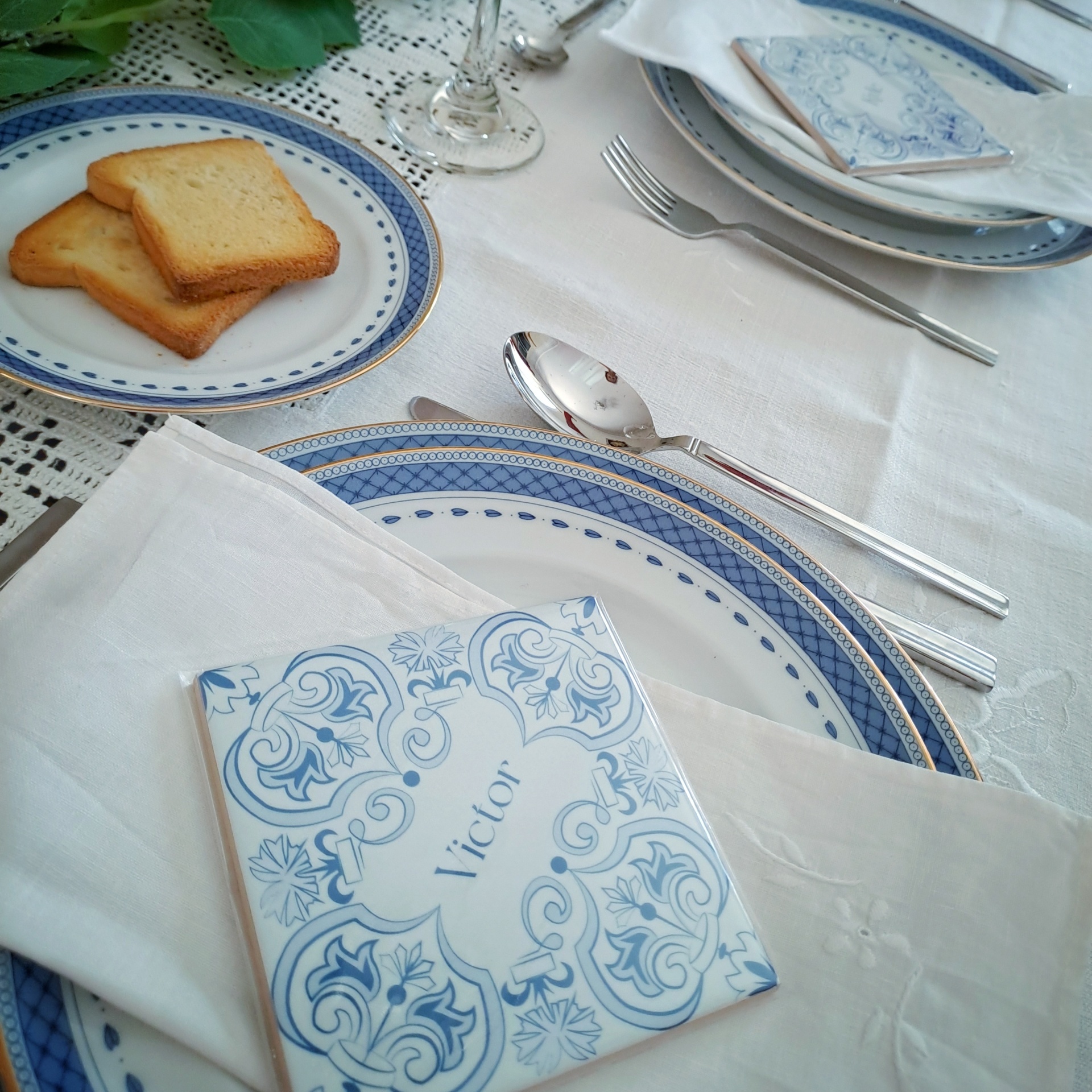 Mesa posta com pratos azuis e brancos, talheres, copo, tostas e guardanapo com azulejo 'Victor'