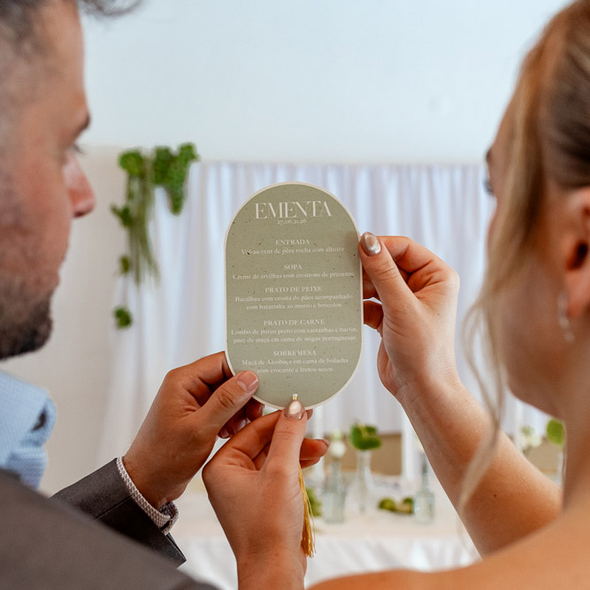 Cartão de ementa oval bege com texto em branco e dourado, segurado por duas pessoas