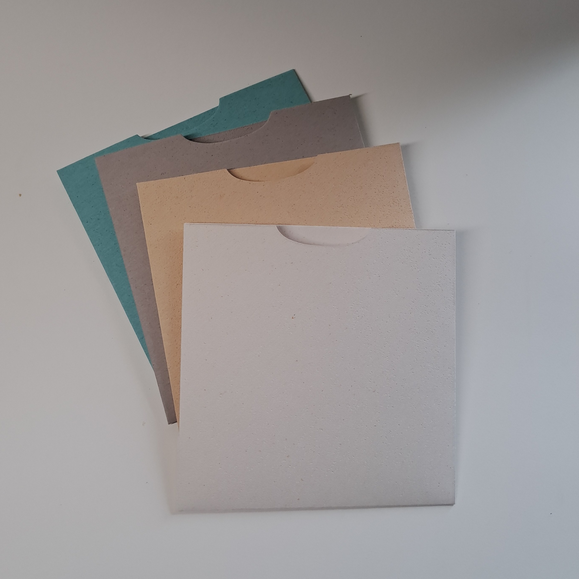 Envelope Cork QD14,5 Luva Conjunto de quatro envelopes quadrados coloridos sobre mesa branca