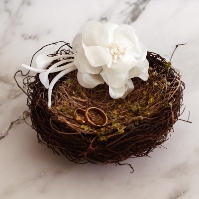 Cesta decorativa com flor branca artificial e dois anéis dourados sobre uma superfície de mármore branca