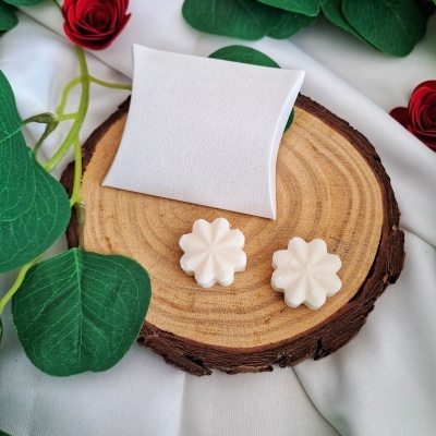 Dois objetos em forma de flor bege clara sobre disco de madeira com embalagem branca, folhas e rosas artificiais