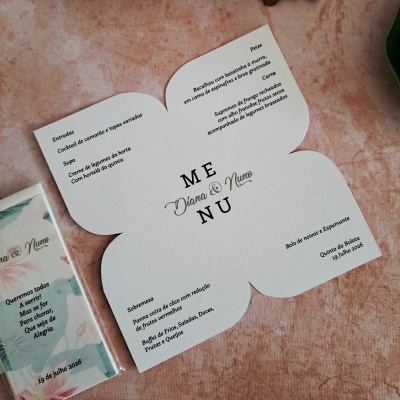 Menu de casamento impresso em papel branco com texto em preto e dourado, nomes Diana & Nuno e data 19 de julho 2026, sobre superfície rosa clara