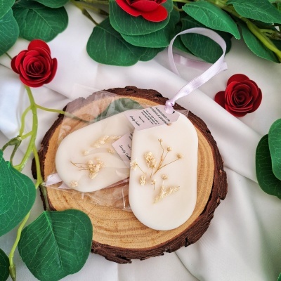 Dois sabonetes decorativos ovais brancos com flores secas embutidas sobre base de madeira com decoração de folhas e flores.