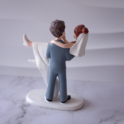 Figura decorativa de casal de noivos em cima de base branca, noivo de fato cinzento a segurar noiva de vestido branco.