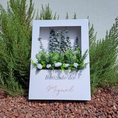 Quadro branco decorativo com plantas artificiais verdes e texto Batizado do Miguel