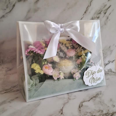 Caixa transparente com laço branco contendo arranjo de flores coloridas e cartão Dia da Mãe
