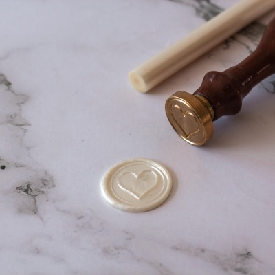 Selo de cera branco com coração, carimbo de metal com cabo de madeira castanha e bastão de cera branca sobre mármore cinzento