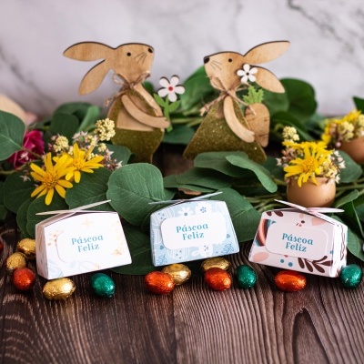 Caixas de papel com texto Páscoa Feliz, ovos de chocolate coloridos, flores e coelhos de madeira num fundo de madeira escura