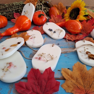 Velas decorativas brancas com flores secas, folhas de outono e abóboras pequenas laranja sobre superfície azul.