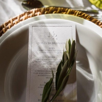 Conjunto de jantar com prato branco sobre prato de vime, menu de papel e rama verde