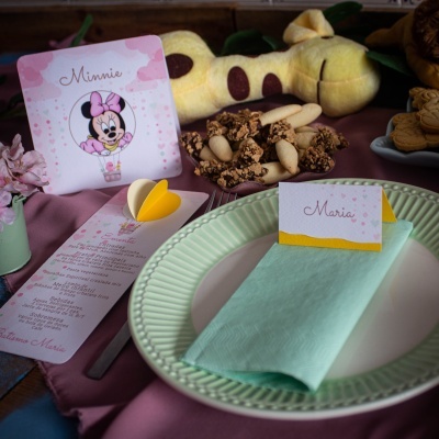 Mesa posta com prato branco e verde, guardanapo azul, cartões com nomes 'Maria' e 'Minnie' e doces ao fundo