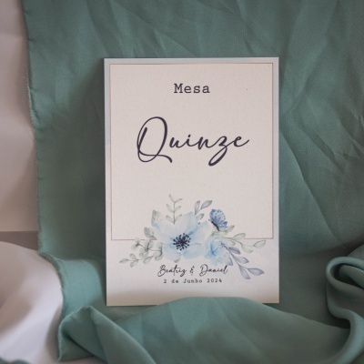 Cartão de mesa branco com design floral azul e texto em português sobre tecido verde-azulado
