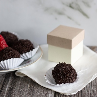 Brigadeiros com granulados de chocolate escuro em forminhas brancas, e caixa bege e branca ao fundo
