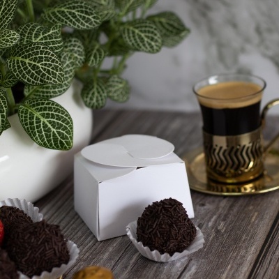 Doces brigadeiros com granulado, caixa branca, chávena de café em suporte dourado, vaso com planta