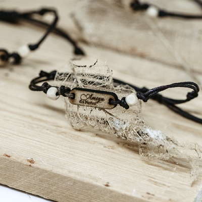 Pulseira de cordão preto com placa de madeira gravada e contas brancas sobre superfície de madeira clara e tecido rendado