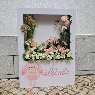 Moldura decorativa branca com flores artificiais e texto 'BATISMO Leonor'