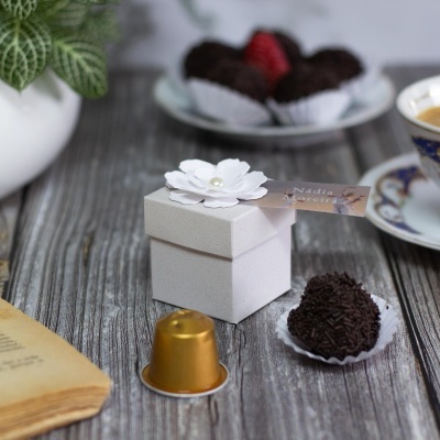 Caixa pequena branca decorada com flor e etiqueta junto a brigadeiro, cápsula de café, livro aberto e chávena de café numa mesa de madeira