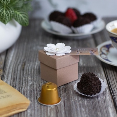 Caixa castanha com flor branca, bombons, cápsula dourada e chávena numa mesa de madeira