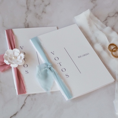 Dois cadernos brancos de votos de casamento com fitas coloridas, anéis dourados e tecido branco sobre mármore.