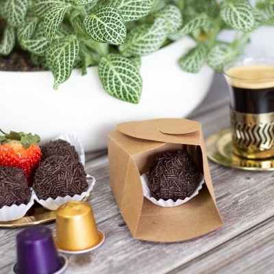 Doces brigadeiros com granulado de chocolate, morango, caixa de papel castanha, cápsulas de café e chávena de café espresso sobre mesa de madeira.