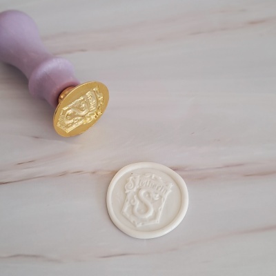 Carimbo dourado com cabo lilás e selo de cera branco com selo Slytherin em relevo