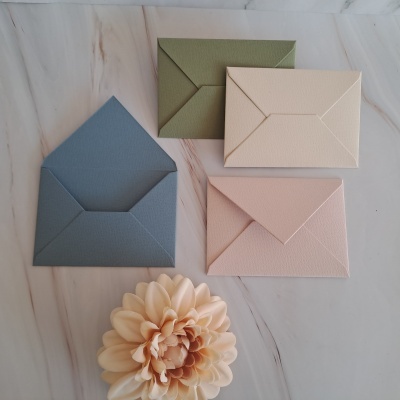 Quatro envelopes coloridos em cartão com textura e uma flor artificial sobre superfície de mármore