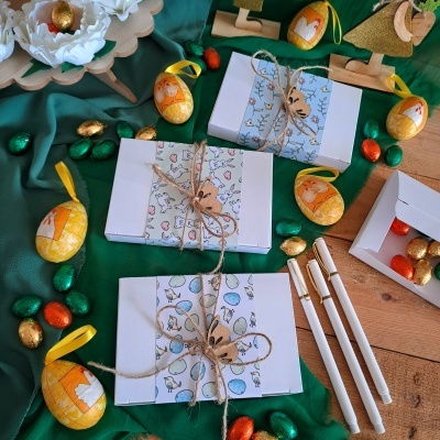 Caixas brancas decoradas com coelhinhos, fitas de corda e ovos de chocolate coloridos sobre tecido verde e madeira.
