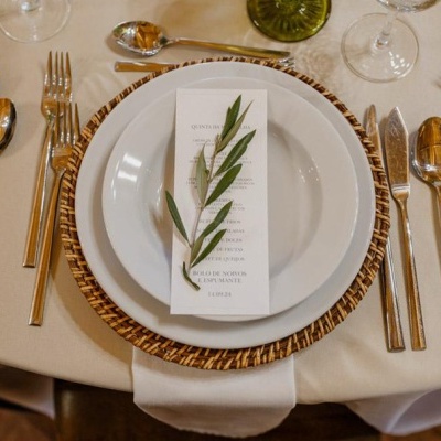 Prato branco com base de fibra natural e ementa decorada com folhas verdes, talheres dourados e copos em mesa com toalha bege