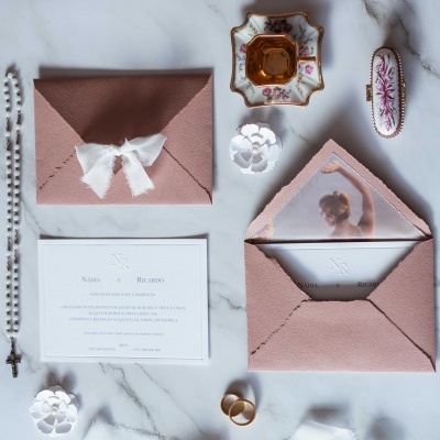 Convite de casamento com envelope rosa, fita branca e acessórios sobre mesa de mármore