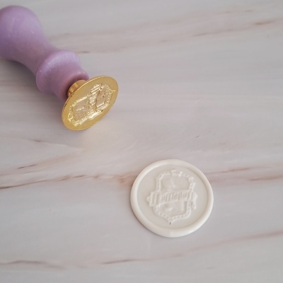 Selo de lacre com cabo roxo, ponta dourada e cera branca com escudo gravado.