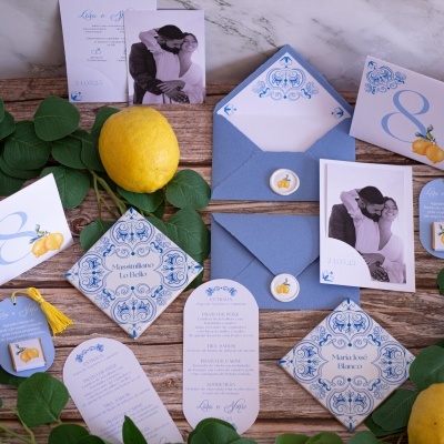 Convites e cartões de casamento azuis e brancos com padrões e limões, acompanhados de fotos a preto e branco sobre mesa de madeira com limões e folhas.