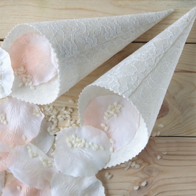 Dois cones decorativos em tecido rendado branco com pétalas de rosa e grãos pequenos brancos