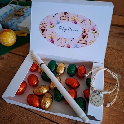 Caixa aberta com ovos de chocolate coloridos, caneta branca e pulseira com pendente de pomba