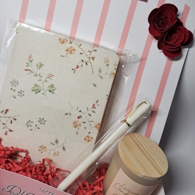 Conjunto de presente com caderno floral, caneta branca com detalhes dourados e vela com tampa de madeira sobre fundo riscado cor-de-rosa e branco