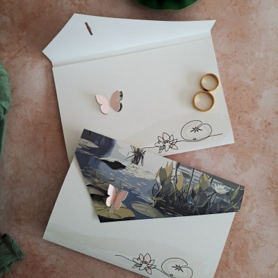 Convite em papel branco com detalhes florais, borboletas cor-de-rosa e envelope com imagem de plantas aquáticas sobre fundo rosa com tecido verde e dois anéis dourados.