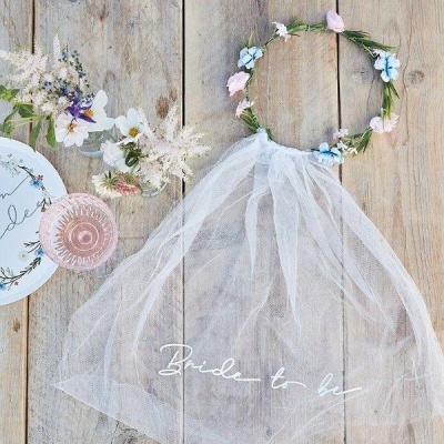 Véu branco com texto Bride to be, coroa de flores, copo rosa e flores sobre madeira clara